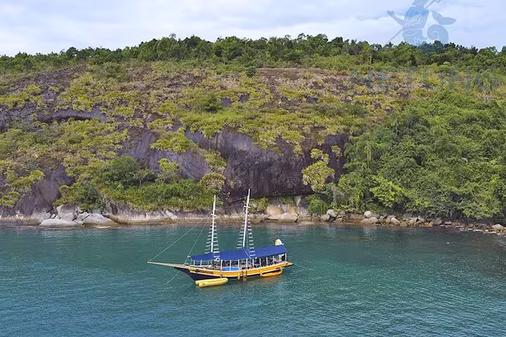 Schooner with paddleboards and kayaks anchored near a verdant cliffside, ideal for water sports enthusiasts.