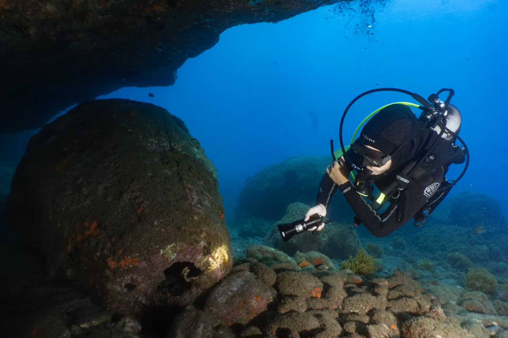 Scuba diver illuminating underwater rock formations on a house-reef dive from shore, highlighting marine exploration.