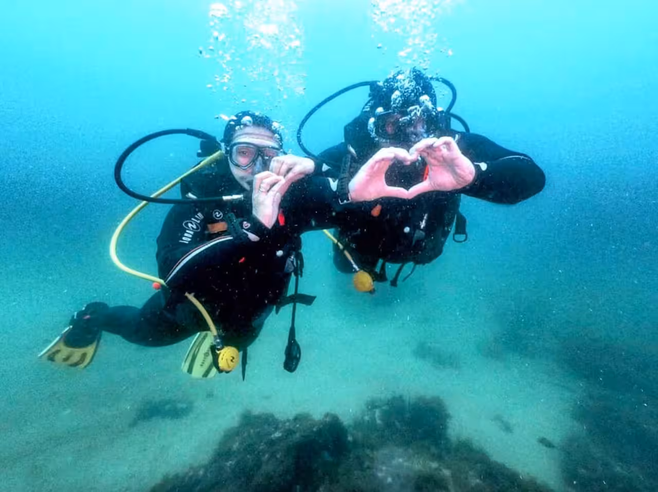 Scuba diving couple forming heart hands underwater on 1 DIVE tour, bubbles rising over rocky seabed