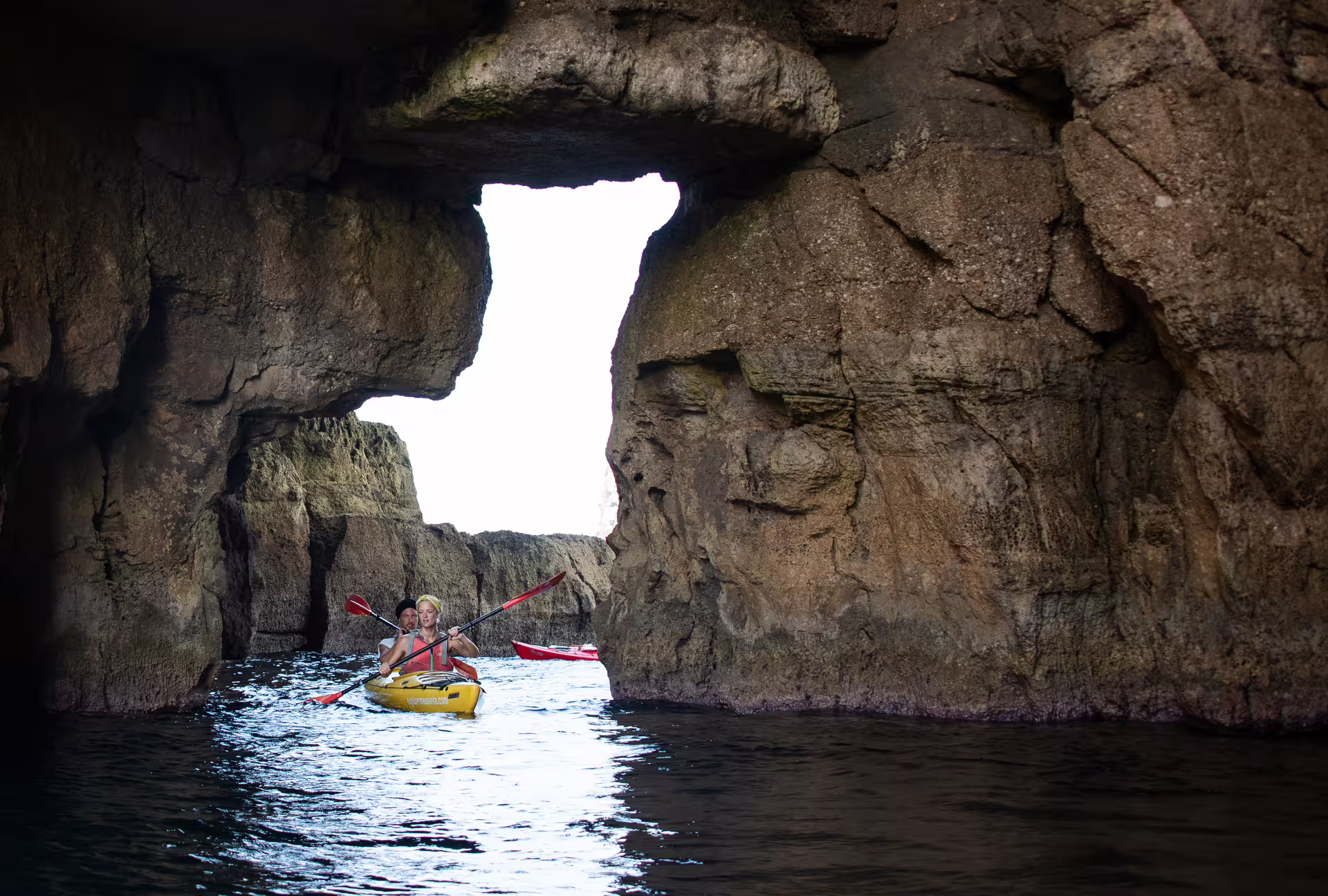 Couple paddling a sea kayak through a sea cave arch in Navarino Bay, Pylos, Greece coastal kayak adventure