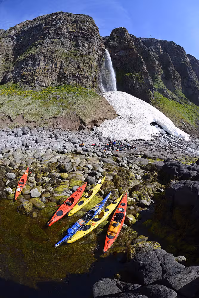 Sea kayaks on a rocky shore below a waterfall and snowfield, showcasing Paddle in the Wild adventure