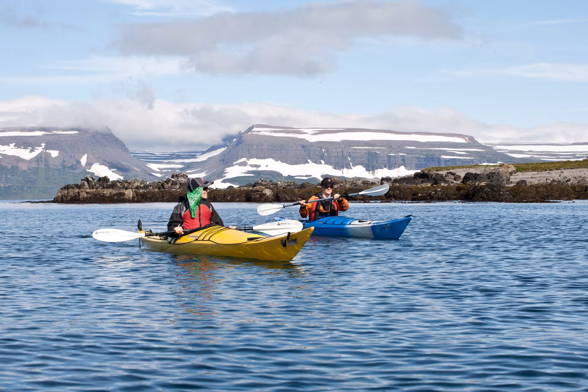 Two guests sea kayaking on Seabird Discovery tour, paddling a scenic fjord with snowy peaks and rocky shoreline