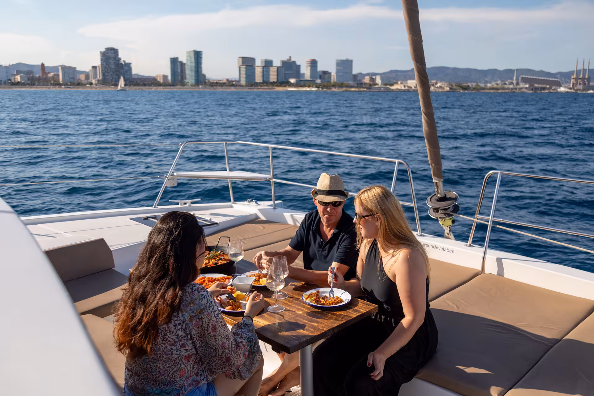 Guests enjoy seafood paella and wine on a sailing yacht with Barcelona skyline, part of a Paella Masterclass