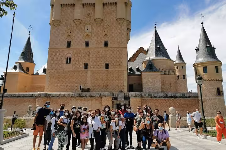 Tour group posing happily outside the historic Alcazar of Segovia under a clear blue sky.