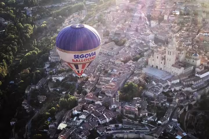 Hot air balloon with Segovia Balloons logo soars above historic Segovia, offering stunning aerial views of the cityscape.