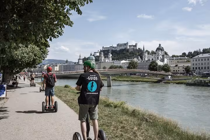 Segway tour group glides along Salzburg's riverside path with a scenic view of the historic fortress.