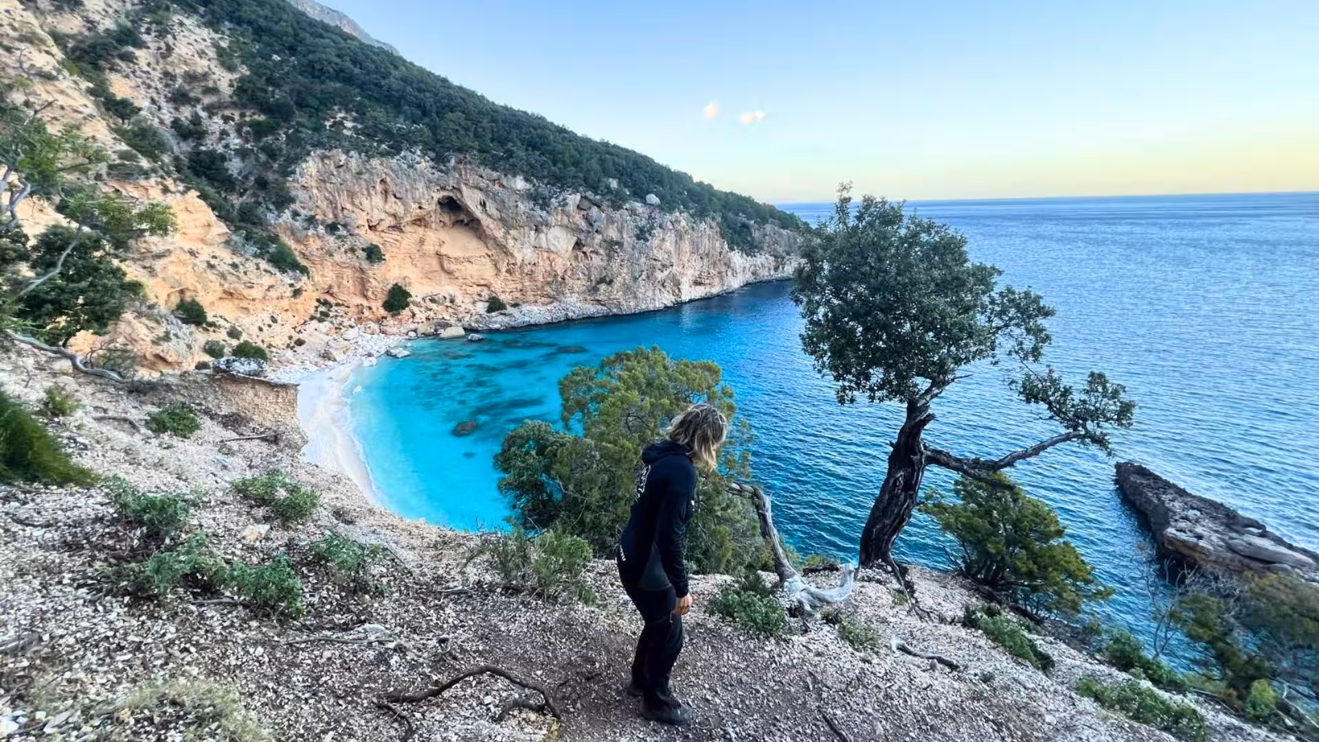 Hiker overlooking the stunning turquoise waters and rugged cliffs of Selvaggio Blu from a cliffside viewpoint.