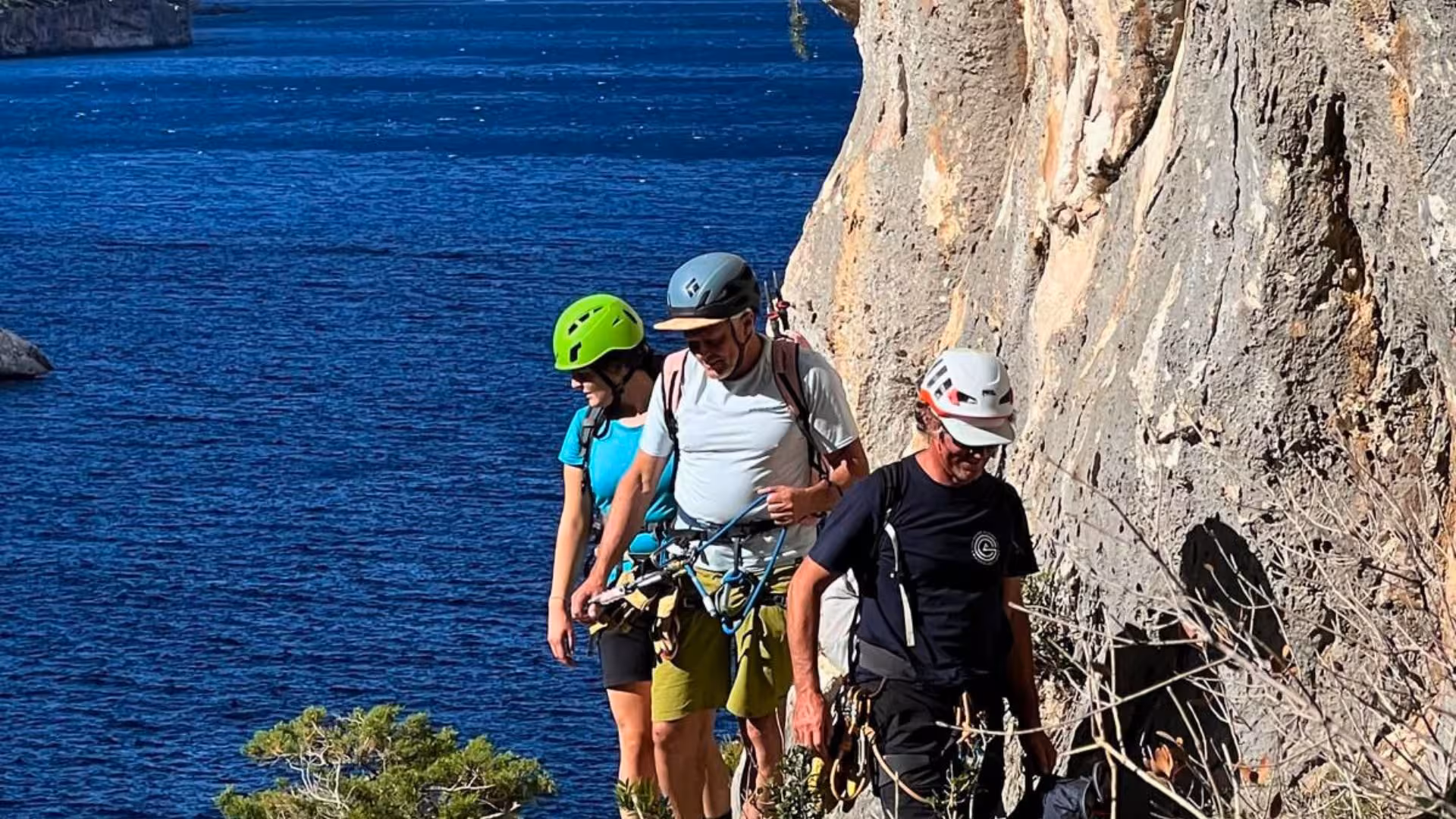 Three climbers with helmets and gear navigating a steep cliff edge on the Selvaggio Blu trek overlooking the sea.