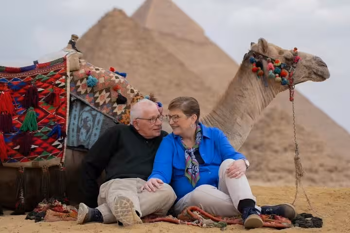 Senior couple sitting by decorated camel at Giza Pyramids during 3-day Cairo tour with museum and free hostel