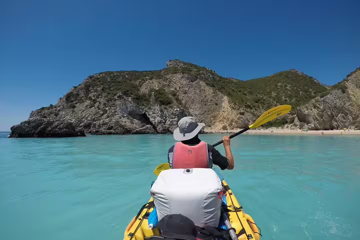 Kayaker paddling through turquoise waters towards rocky cliffs in Sesimbra's Arrábida Natural Park on a sunny day.