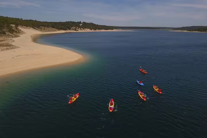 Kayakers paddle on a serene lagoon near Sesimbra, Portugal, surrounded by sandy shores and lush greenery under a clear sky.