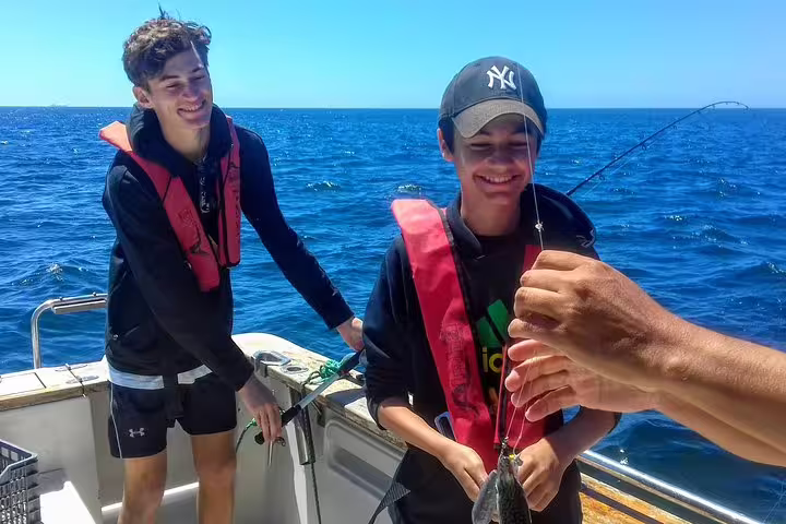 Two smiling anglers enjoy a successful catch on a sunny Sesimbra sport fishing tour, wearing life jackets on a boat.