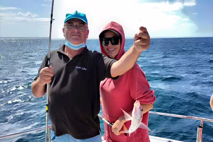 Two happy anglers on a sport fishing tour in Sesimbra hold up a fresh catch against a backdrop of the sparkling ocean.