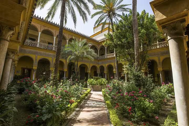 Ornate yellow courtyard with arched galleries, palm trees, and colorful flower gardens in Seville, Spain