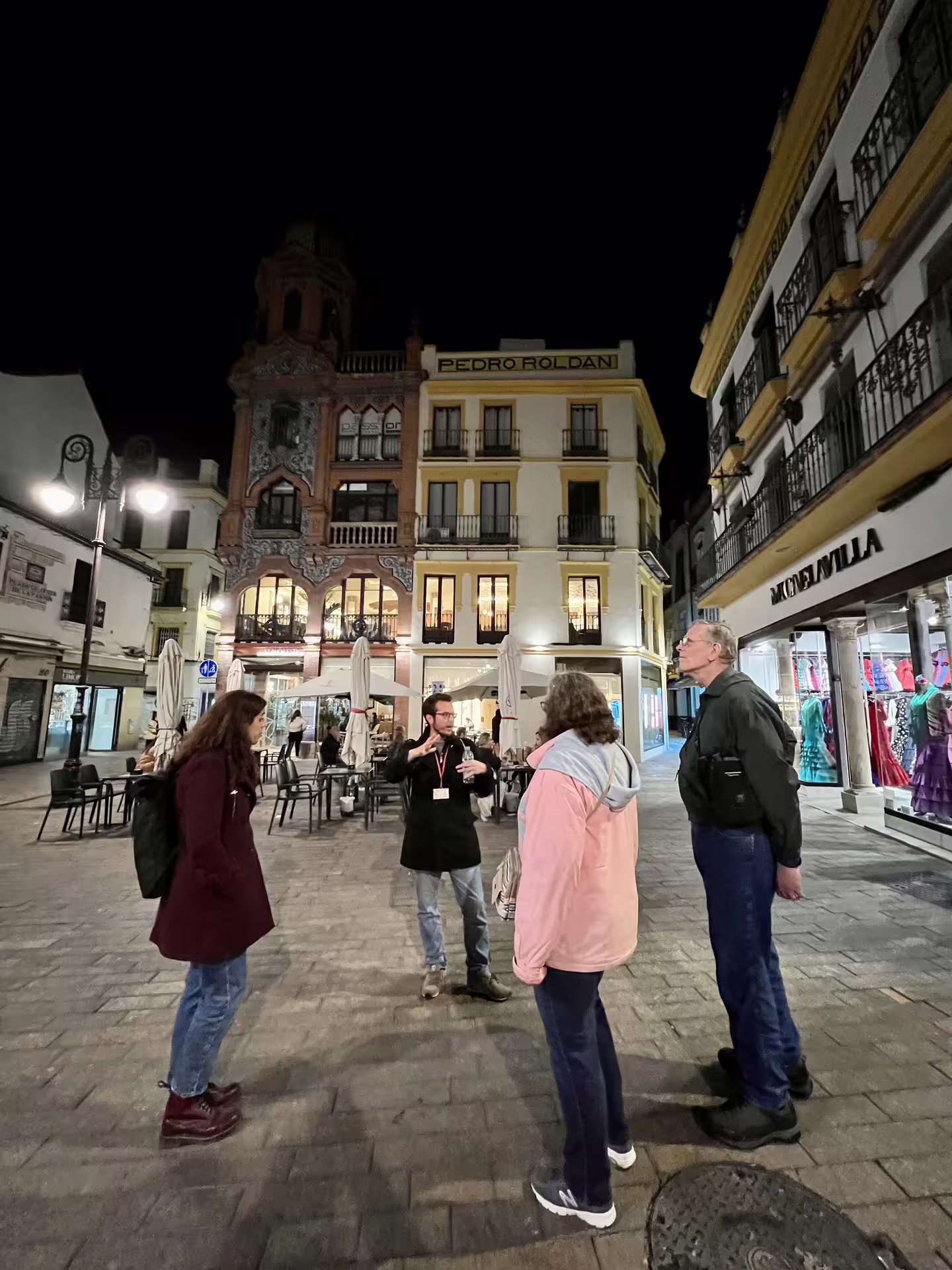 Tour guide leading a group on a nighttime Seville Mysterious and Paranormal Tour in a historic plaza under streetlights.