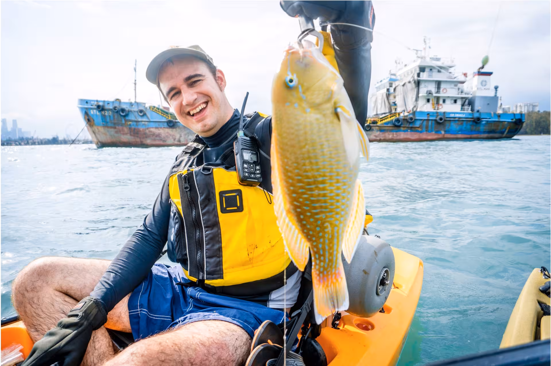 Smiling kayaker holding a vibrant fish with a ship in the background at Shipwreck Bay Marine East fishing tour.