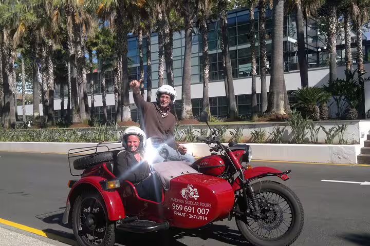 Two people enjoy a thrilling sidecar adventure in sunny Funchal, showcasing Downtown Delights tour with palm trees and modern architecture.