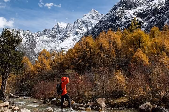 Hiker in red jacket explores the scenic autumn landscape of Siguniang Mountain with snow-capped peaks in the background.