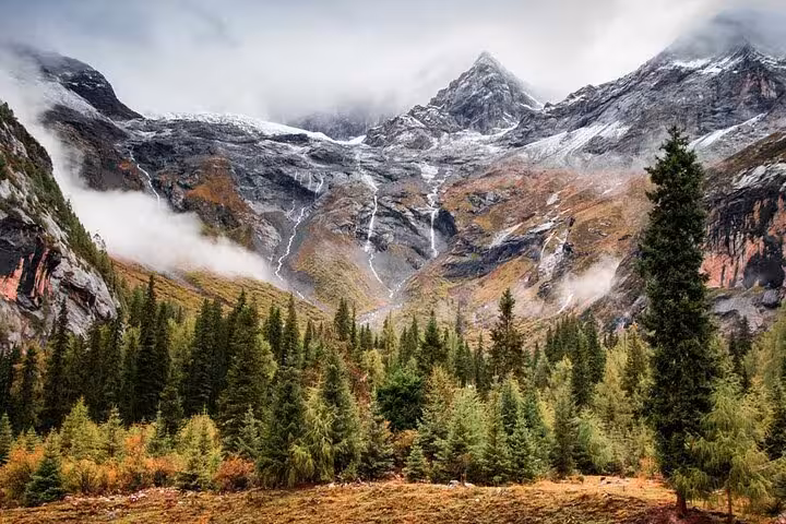 Scenic view of Siguniang Mountain's rugged peaks shrouded in mist, surrounded by lush green forest and autumn colors.