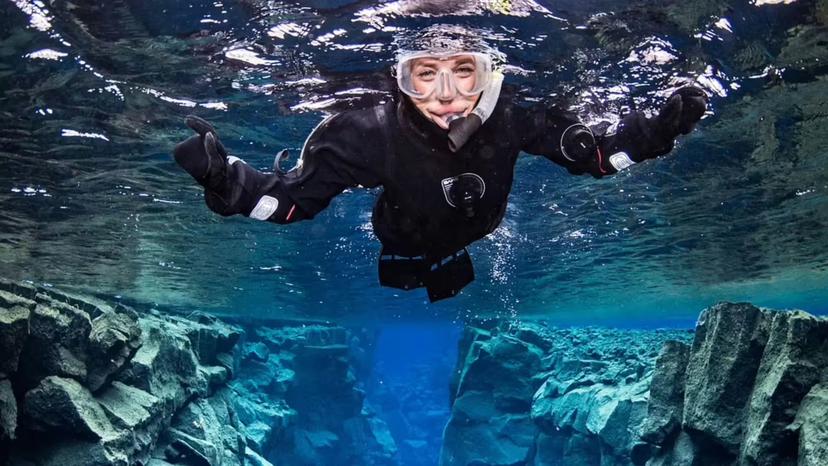 Smiling snorkeler in drysuit floating underwater above rocky Silfra fissure, Iceland