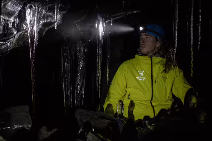 Adventurer in a bright yellow jacket explores the dark, icy Leidarendi Lava Tunnel, illuminated by headlamp light.