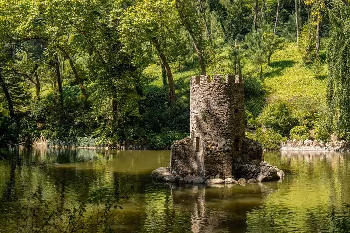 Scenic view of a small stone tower surrounded by a serene pond and lush forest in the gardens of Sintra's Pena Park.