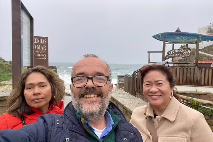 Three tourists smiling near the ocean at Azenhas do Mar, a scenic stop on a private Sintra tour from Lisbon to Pena Palace.