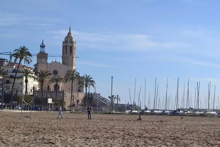 Scenic view of Sitges beach with palm trees and historic church, perfect for a cultural and culinary tour.