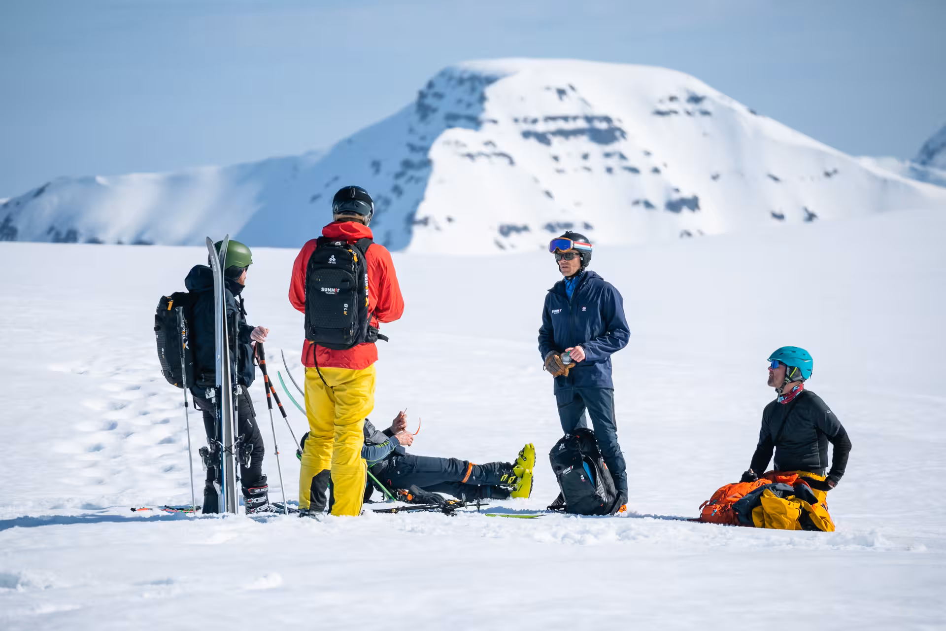 Group of skiers resting on snow-covered mountain during heli-tour adventure with stunning snowy peaks in the background.
