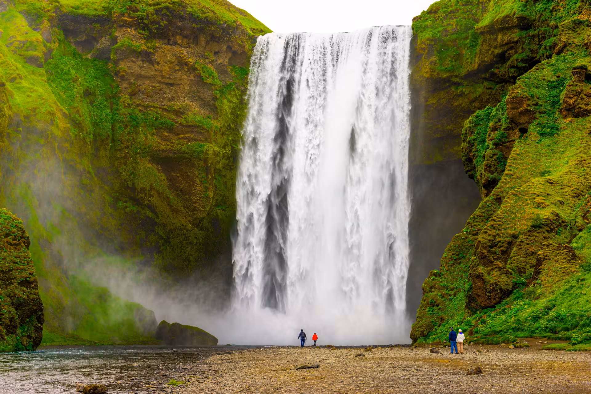 Visitors admire the majestic Skógafoss waterfall surrounded by lush green cliffs on a glacier tour in Iceland.
