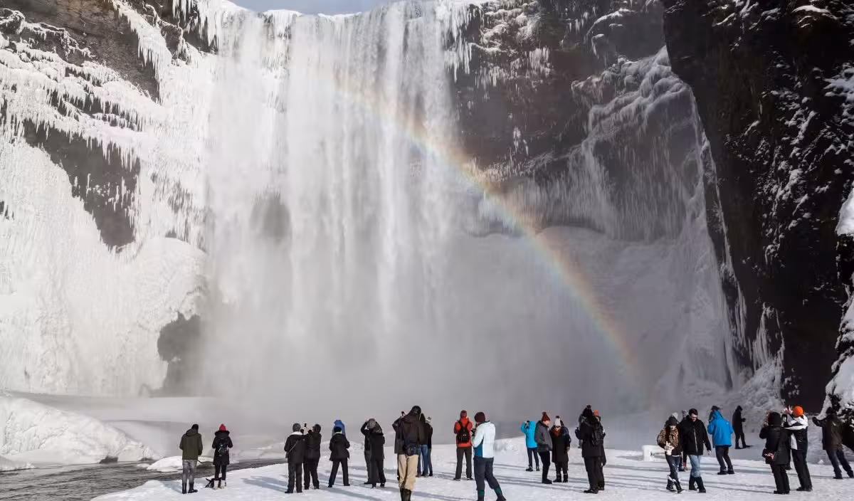 Tourists admire Skógafoss waterfall with icy backdrop and rainbow on Iceland stopover tour.