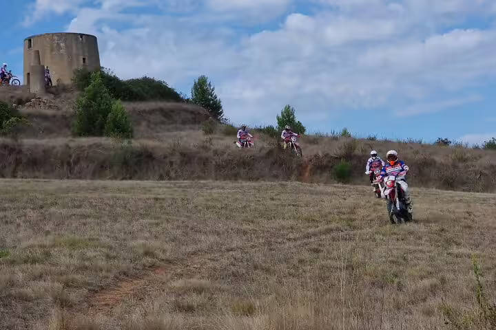 Small group of enduro riders navigating rugged Lisbon terrain near an ancient stone tower under a blue sky.