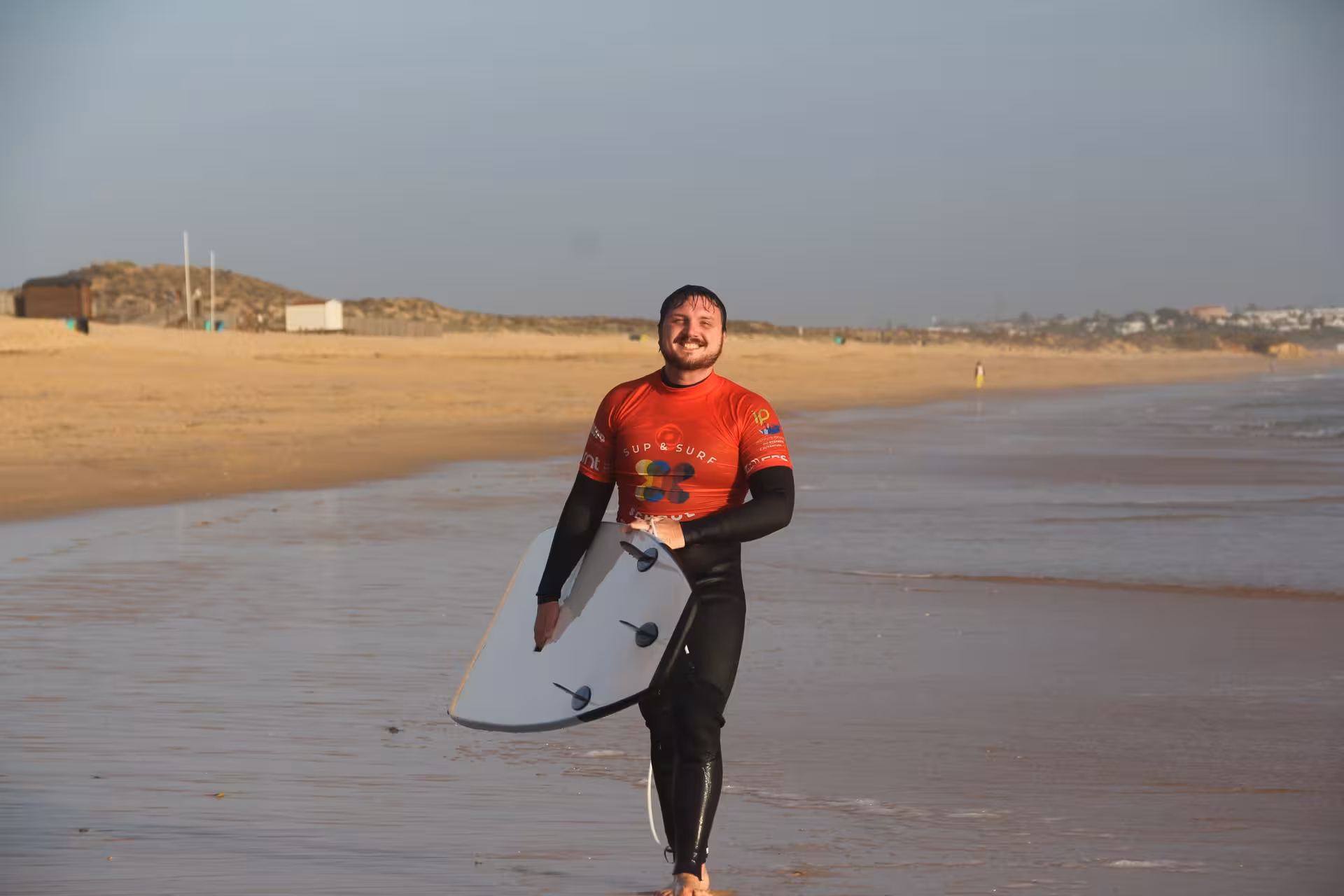 Smiling surfer in red rash vest carrying a board along a wide sandy beach after a fun surf lesson