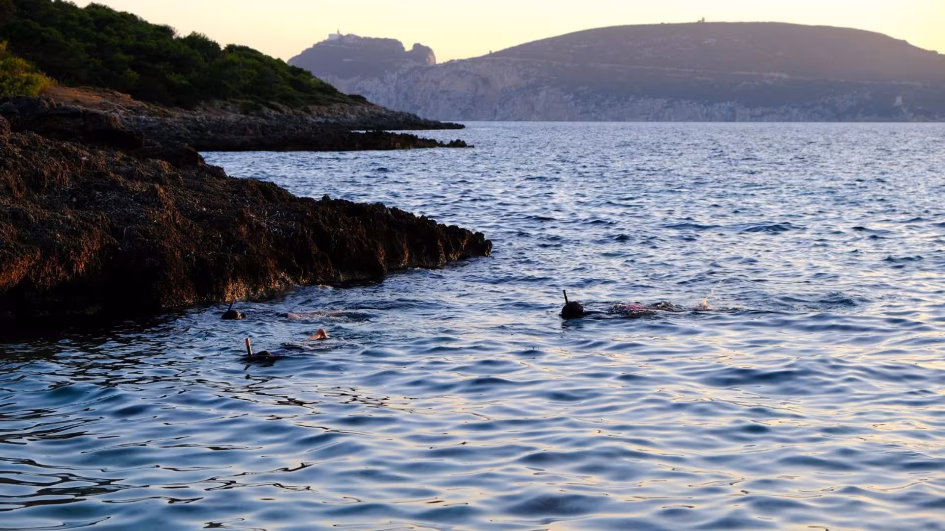 Snorkelers exploring the clear blue waters near rocky shores in Porto Conte park, Alghero at sunset.