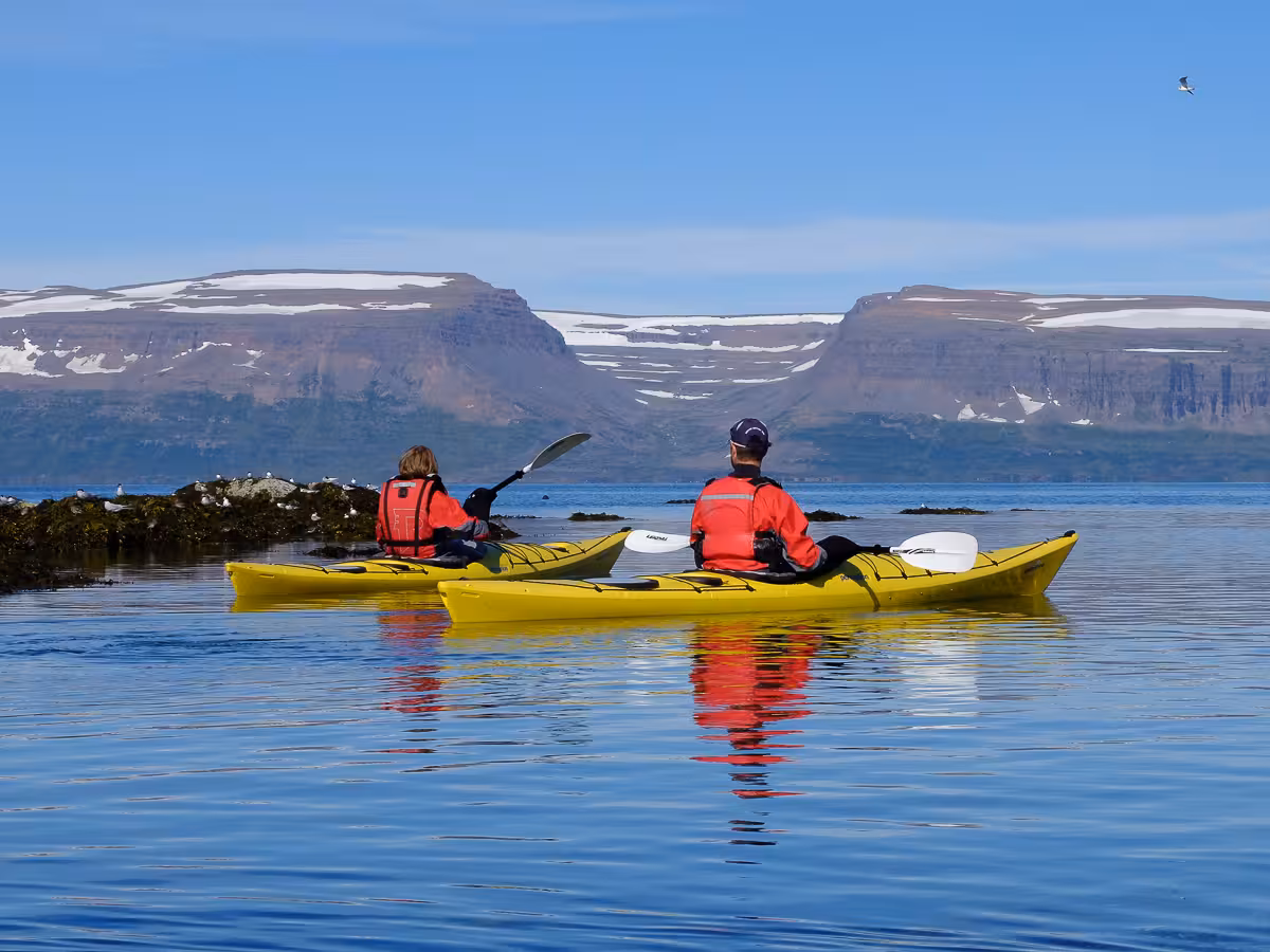 Two kayakers in red vests glide across Arctic fjord with snowcapped mountains on Solar Eclipse Wild Camp Discovery