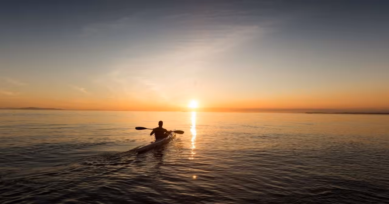 Solo kayaker paddles on calm sea at sunset, capturing serene outdoor adventure.