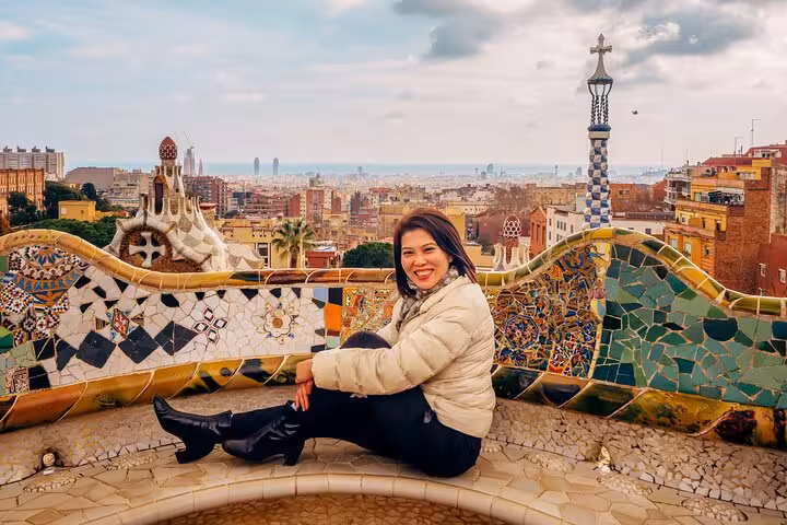 Solo traveler posing on Park Güell mosaic bench with panoramic Barcelona skyline, Gaudí architecture, and Mediterranean Sea views.