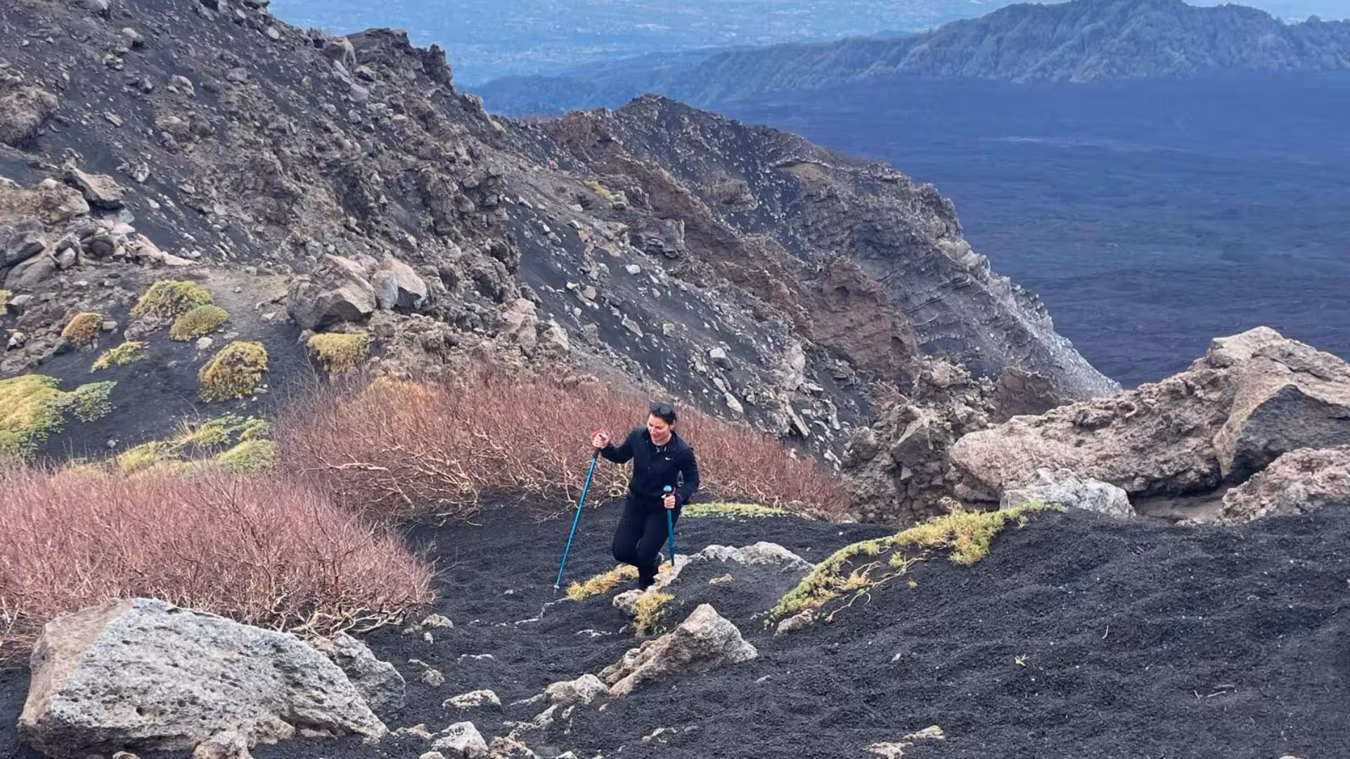 Solo trekker navigates rugged volcanic landscape on Etna, surrounded by rocky cliffs and sparse vegetation.