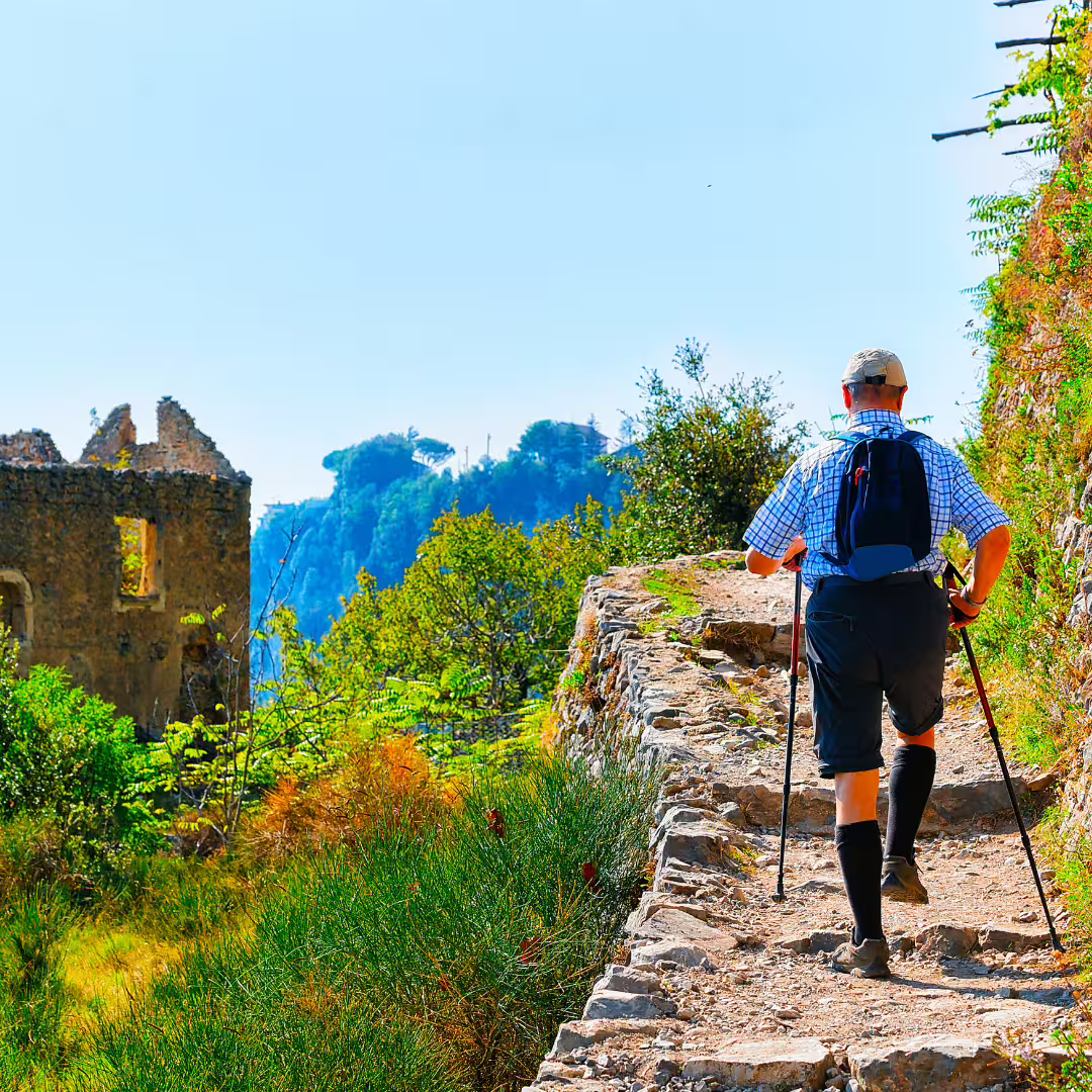 Solo trekker with poles climbs rocky Path of the Gods past ancient stone ruins and lush Mediterranean vegetation on the Amalfi Coast