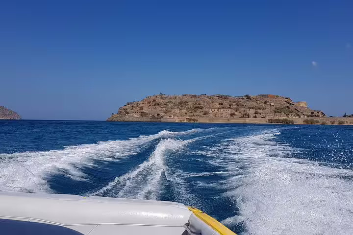 A speedboat leaves a frothy trail as it departs from Island Spinalonga, set against the vibrant blue sea and clear sky.