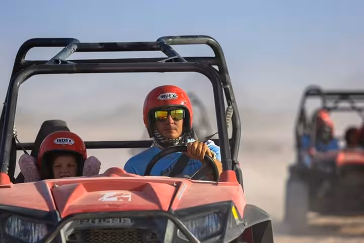 Father and child driving red spider car buggy in Marsa Alam desert safari, with convoy in background