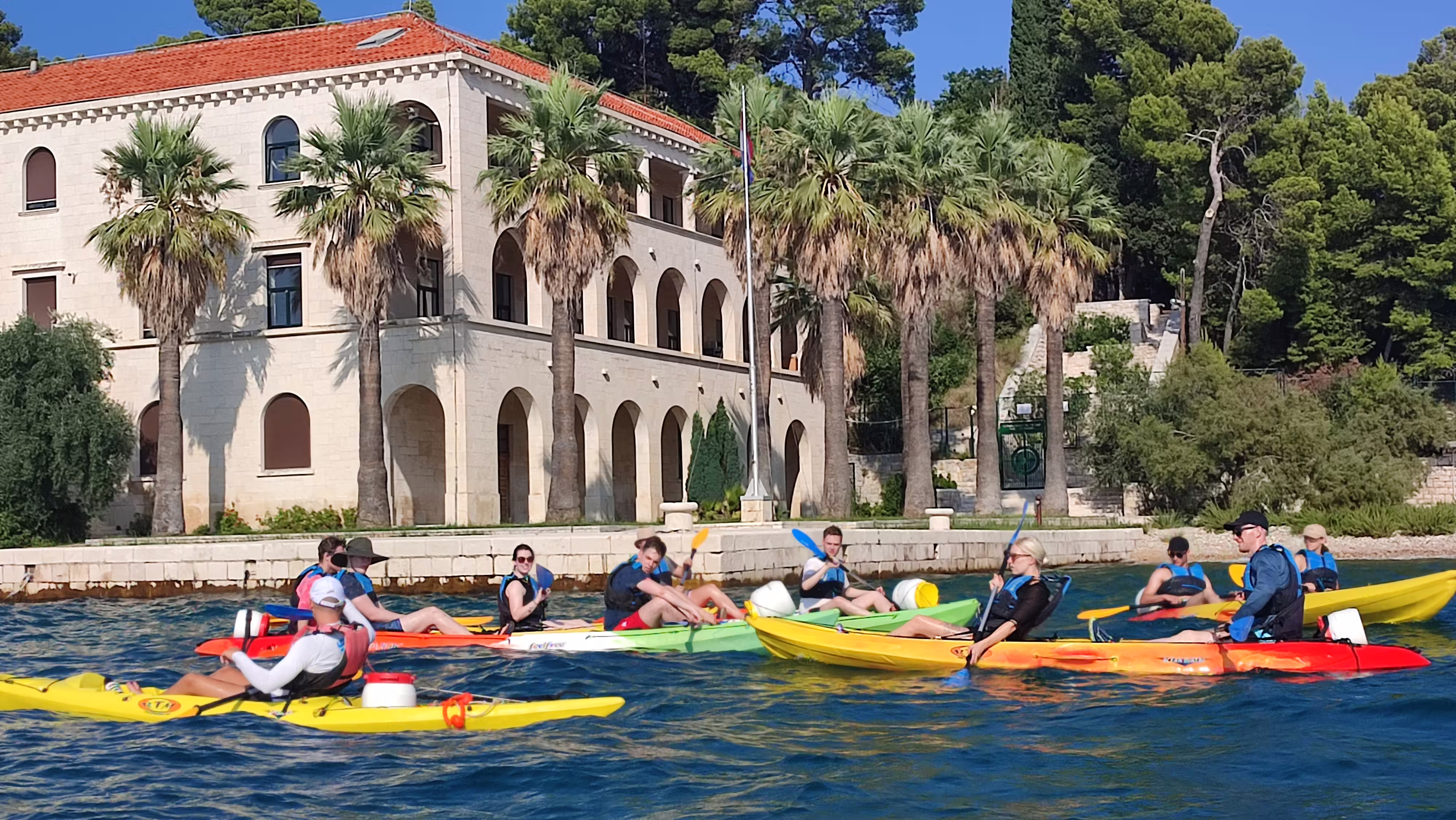 Split sea kayaking tour group paddling by palm-lined villa on the Adriatic, a top kayaking and snorkeling stop