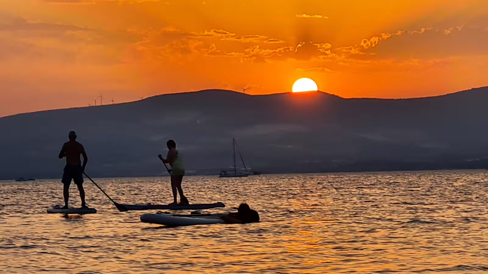 Silhouettes stand up paddleboarding at golden sunset near Split, Adriatic SUP tour with wine and sailboat view