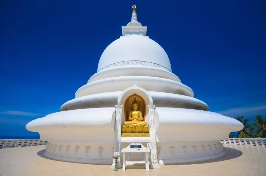 White Peace Pagoda with golden Buddha statue against clear blue sky in Sri Lanka's coastal region.