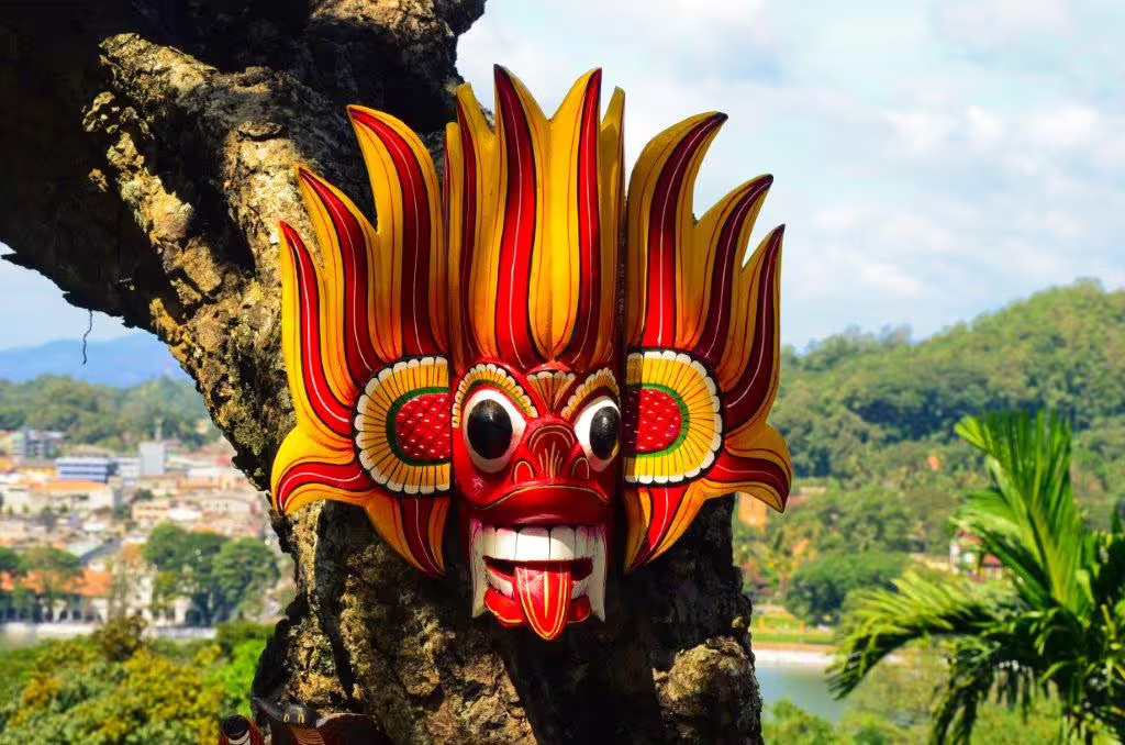 Colorful traditional Sri Lankan mask displayed on a tree with scenic hilly landscape and blue skies in the background.