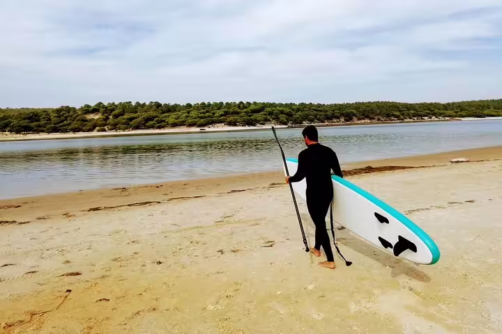 Person in wetsuit carrying paddleboard towards tranquil Lisbon shoreline for stand up paddle adventure experience.