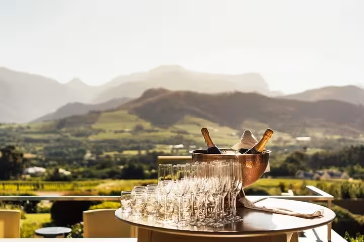 Champagne bottles and glasses on a table with panoramic Stellenbosch vineyard views in the background.