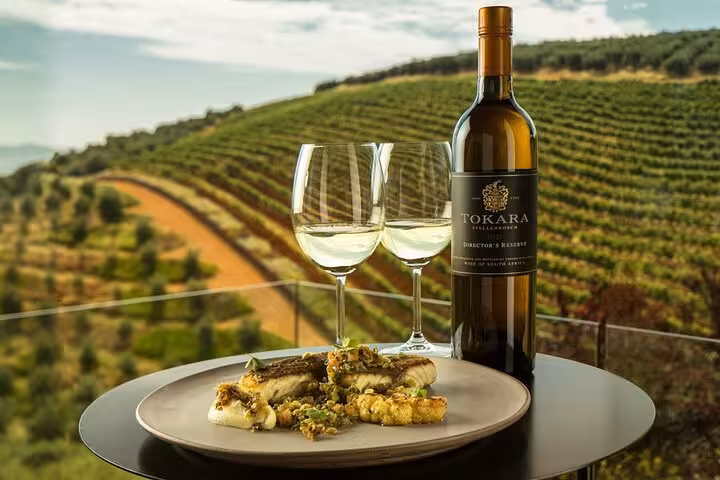 Tokara wine bottle with two glasses and gourmet dish overlooking lush Stellenbosch vineyards.
