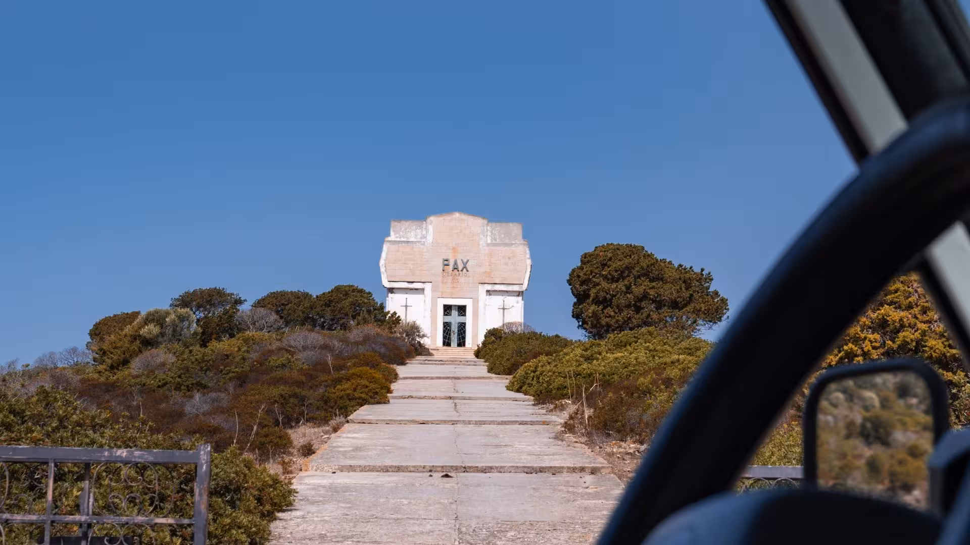 View from minivan of historic building on Asinara Island, surrounded by lush greenery under a clear blue sky.