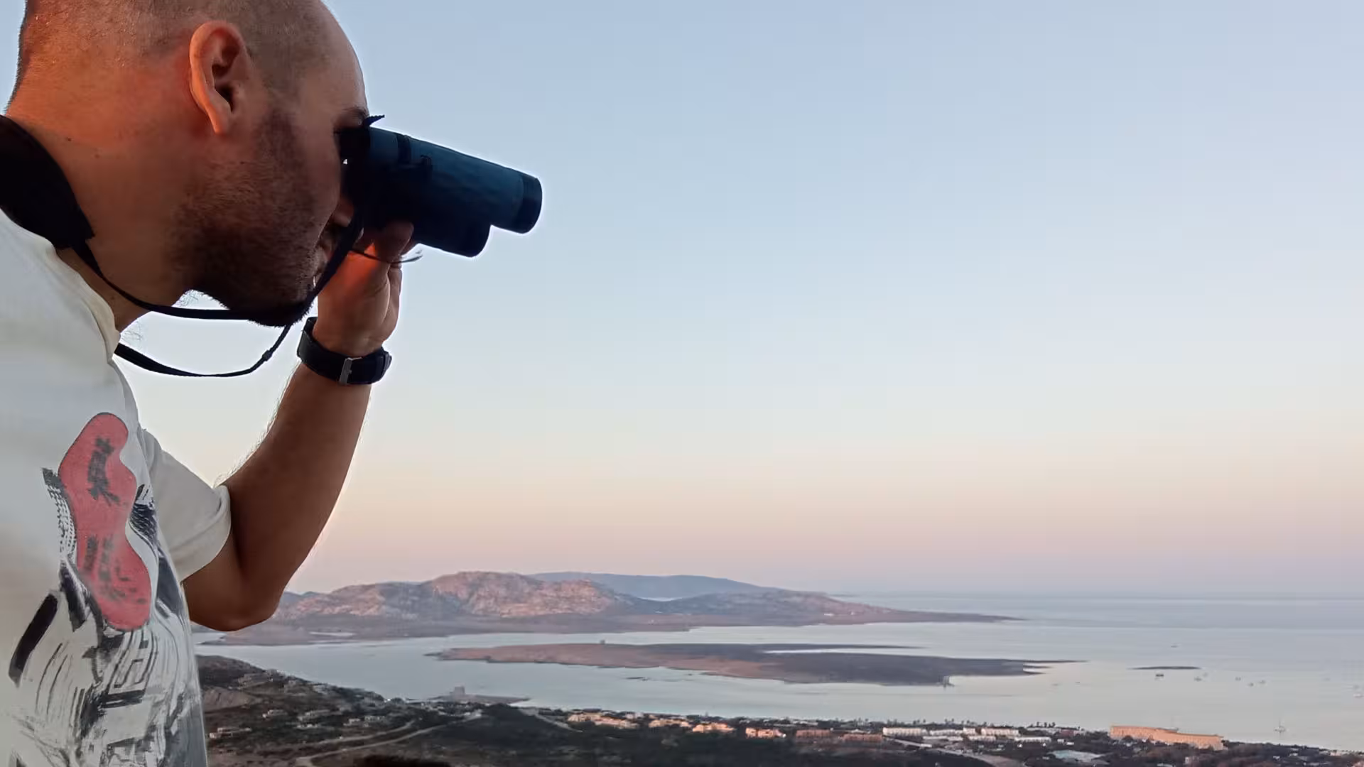 Man with binoculars enjoying sunset view from Capo Falcone during Stintino hike, overlooking the sea and landscape.